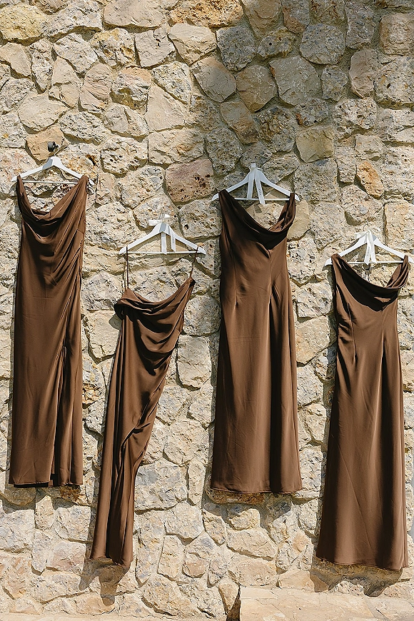 Bridesmaid dresses in brown bridesmaid dresses hang on white hangers, their satin sheen catching sunlight against a stone wall with shadows