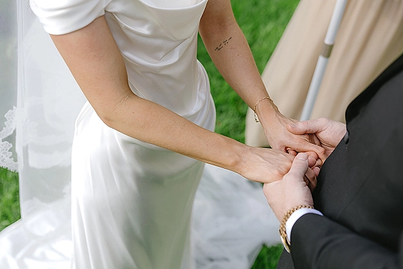 Wedding vows as bride and groom hold hands during ceremony, veil and bracelet visible beside his cuff and wristwatch on grass lawn