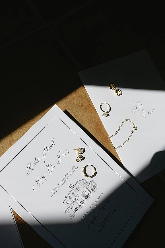 Invitation suite wedding invitation flat lay with calligraphy cards, envelopes, gold bands and engagement ring on brown paper in shadowed light