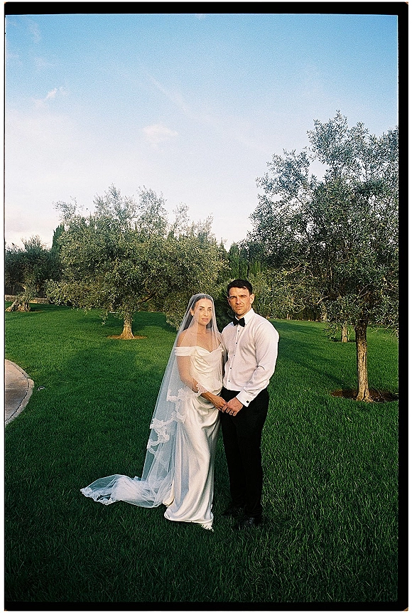 Couple portrait outdoors, bride and groom holding hands, her lace veil flowing behind on a green lawn with trees and blue sky