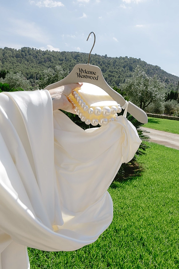 Wedding dress on a wooden hanger with a pearl necklace and off-the-shoulder satin sleeves, held by a hand on a garden path