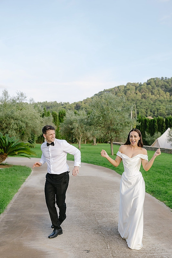 Couple portrait of bride and groom walking, bride in off shoulder wedding dress laughing beside groom in bow tie on a garden path