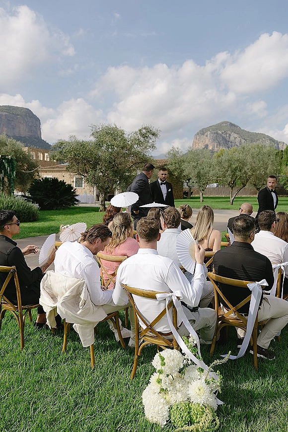 Wedding ceremony with guests seated in wooden chairs, white ribbon bows and paper parasols, facing the groom on a lawn by mountains