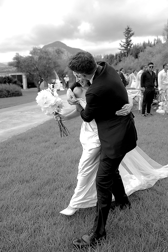 Wedding hug moment as bride and groom embrace laughing, bouquet in hand, on a lawn with mountains, trees, and guests behind