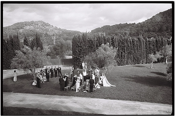 Outdoor wedding ceremony with bride walking down the aisle in a long veil, guests seated on lawn chairs beneath trees and hills