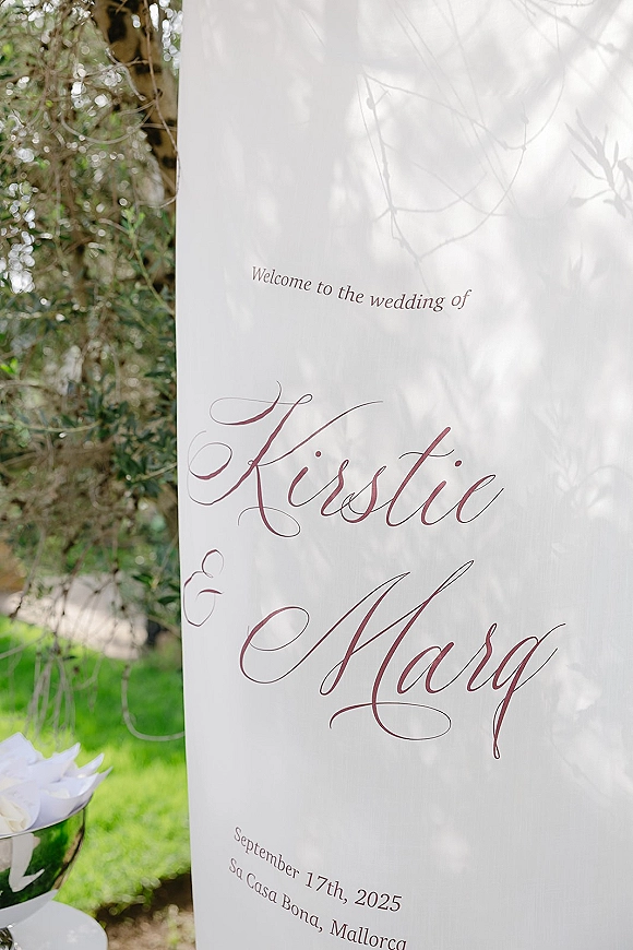 Wedding welcome sign with calligraphy lettering in burgundy on a white board, displayed outdoors on a lawn with greenery and tree branches