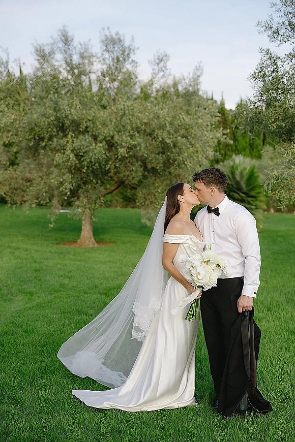 Wedding kiss portrait of bride and groom kissing, bride in off-the-shoulder gown holding white orchid bouquet on lawn by olive trees