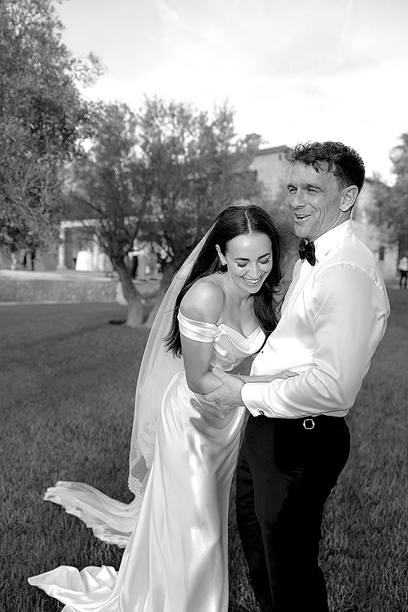 Couple portrait in a black and white wedding portrait as bride and groom laugh, embracing on a lawn with trees and a building behind