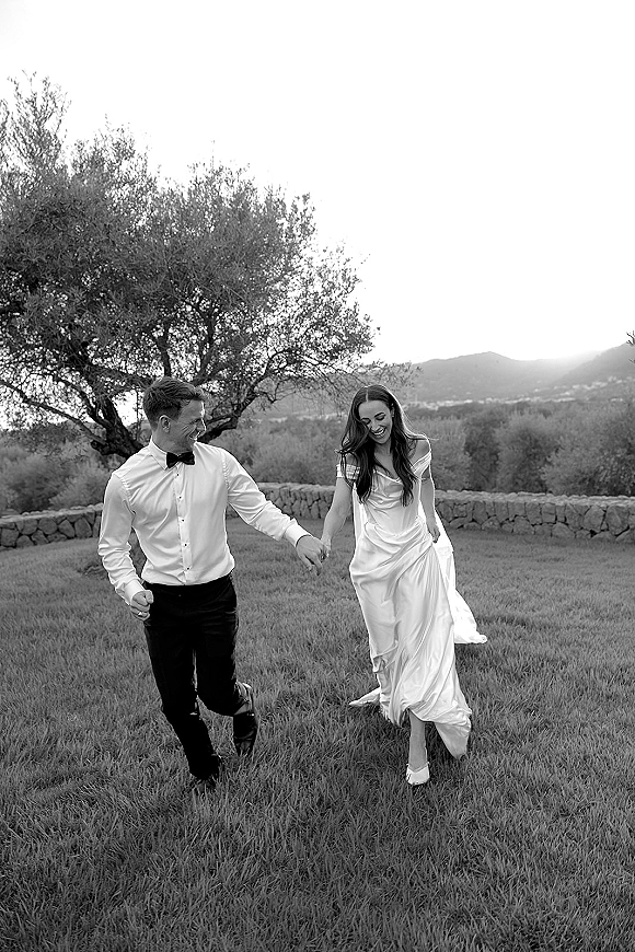 Couple portrait of bride and groom walking hand in hand, bride in off-the-shoulder satin gown and heels on a grassy lawn by a stone wall