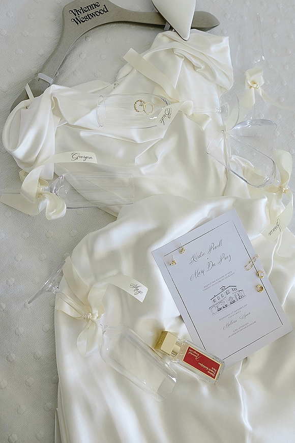 Wedding detail flatlay with wedding invitation suite, rings, earrings, perfume, champagne flutes, ribbons and name tags on white fabric