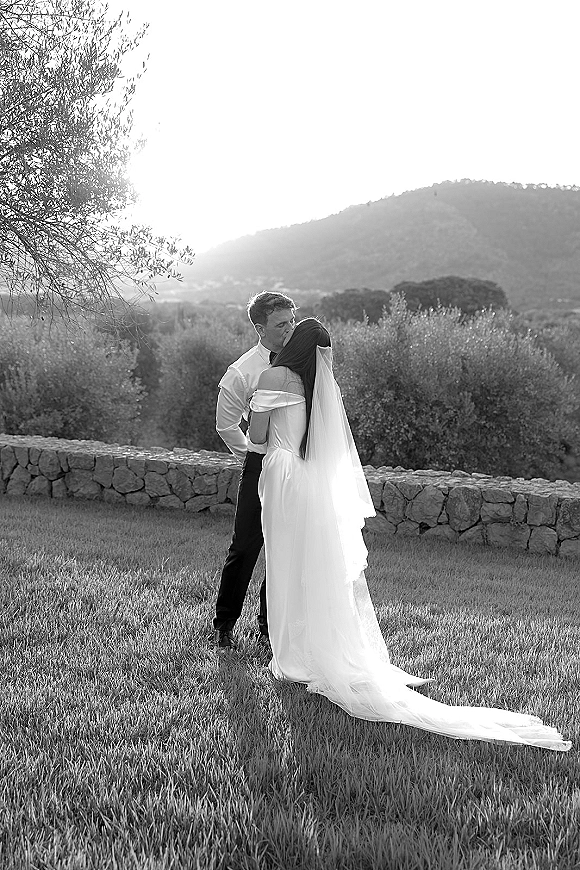 Wedding kiss portrait of bride and groom kissing as her veil lifts, standing by a stone wall with trees and hills beyond