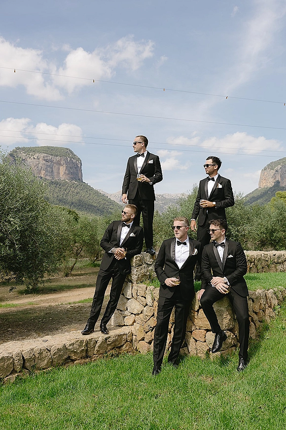 Groomsmen portrait of friends in black tuxedos and sunglasses holding whiskey glasses on a stone wall with mountains and string lights behind