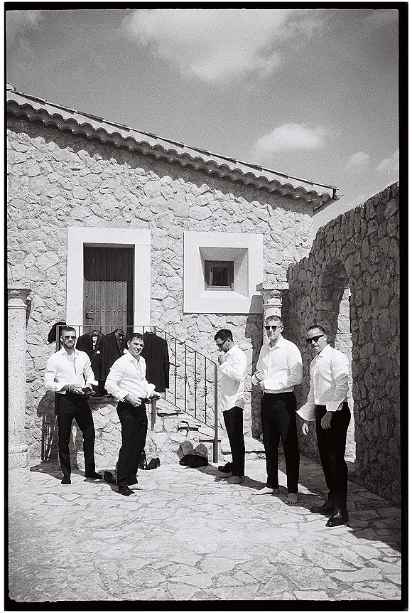 Groomsmen getting ready in white shirts, wearing sunglasses and adjusting outfits on stone steps outside a rustic stone courtyard archway