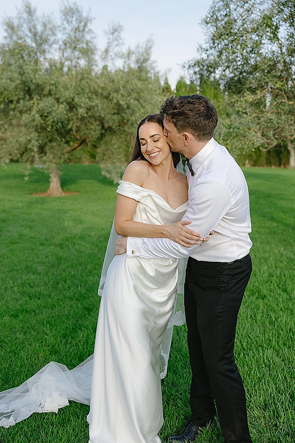 Wedding couple portrait of bride and groom embrace as he kisses her cheek, her off-the-shoulder satin dress and lace veil on a green garden lawn