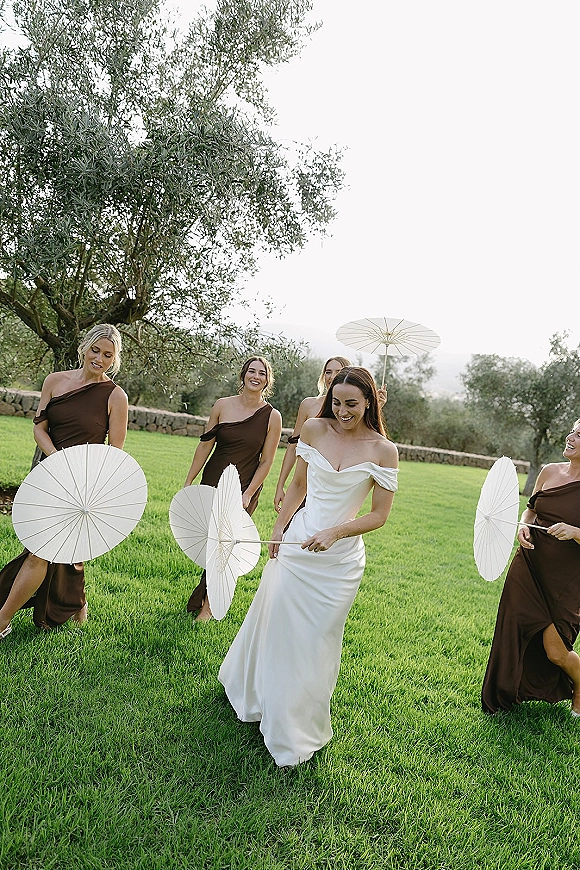 Bridal party photo of the bride with bridesmaids walking on a grass lawn, bridesmaids holding paper parasols near olive trees and stone wall