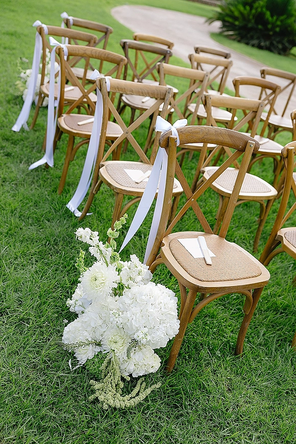 Ceremony seating with outdoor ceremony chairs lined in rows, white ribbon accents and programs, plus a white floral arrangement on a lawn path