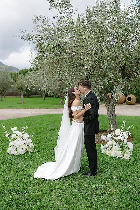 Wedding kiss portrait of bride and groom kissing, her cathedral veil flowing, by white florals along an olive grove garden path under clouds