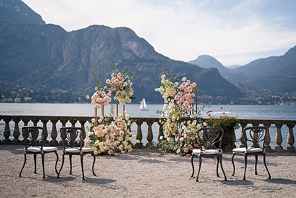 Ceremony backdrop with pastel rose and greenery floral arrangements on pedestal stands, black metal chairs on a terrace overlooking a mountain lake