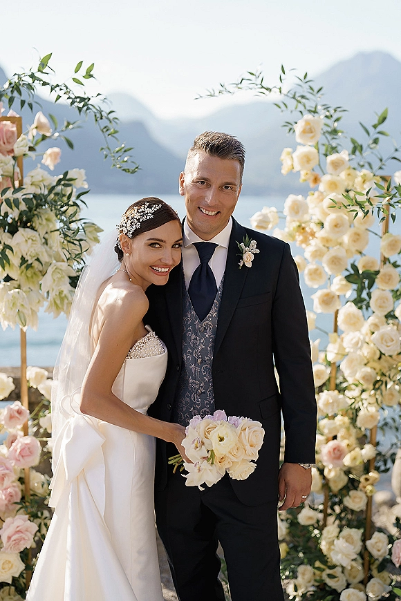 Wedding couple portrait of bride and groom smiling under a floral arch, bride holding white and blush rose bouquet with greenery by a lake and mountains
