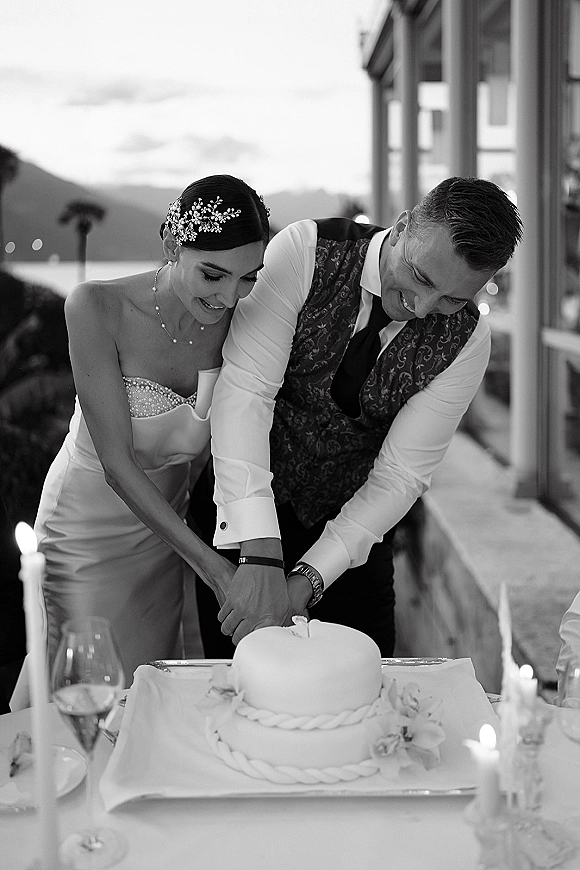 Cake cutting moment as bride and groom guide the knife into a classic white wedding cake on a candlelit terrace by water and mountains at dusk