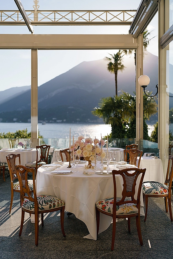 Reception tablescape with round wedding table setup on white linen, pastel rose centerpiece and taper candles on a terrace with lake and mountains