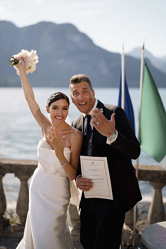 Wedding couple portrait of newlyweds showing rings, bride holding bouquet and marriage certificate on a stone balustrade by a lake and mountains