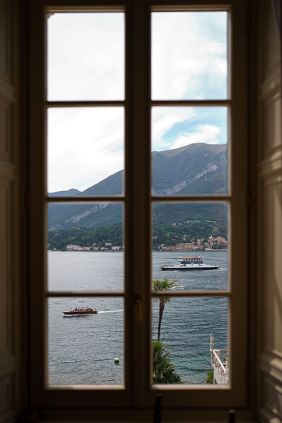 Wedding venue view through a window frame, showing a lake view window with boats, shoreline village, palm tree, and mountains beyond
