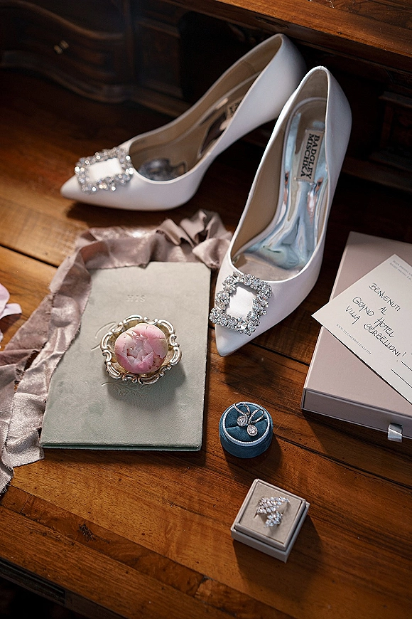 Bridal shoes flatlay with white wedding heels and crystal buckles beside invitation card, rings and earrings on a wooden tabletop