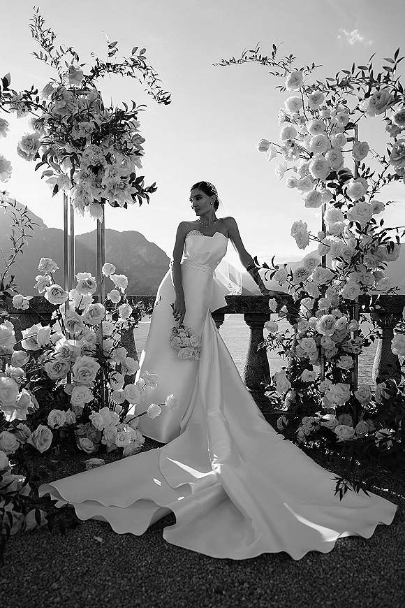 Bridal portrait of a bride in a strapless wedding dress with long train, holding a rose bouquet on a stone terrace by lake and mountains