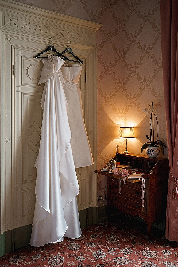 Wedding dress hanging on a hanger, strapless satin gown with oversized bow beside champagne, shoes and bouquet on a vintage desk