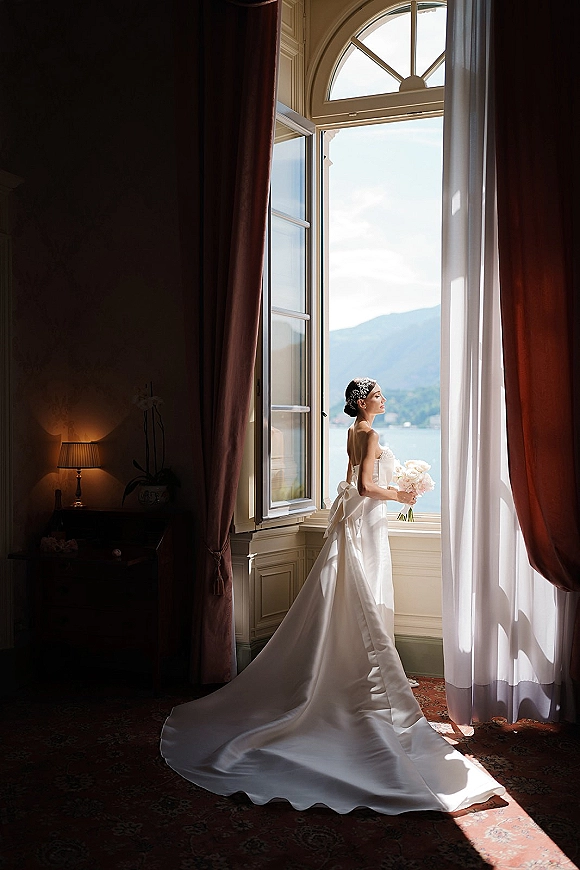 Bridal portrait of a bride by window holding a bouquet in a strapless gown with long train and bow back, framed by an arched window with mountain and lake view