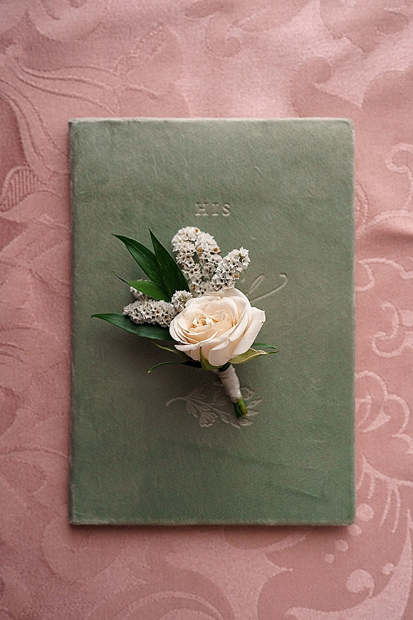 Wedding vows book with embossed cover beside a twine-wrapped white rose boutonniere on pink textured lace fabric backdrop