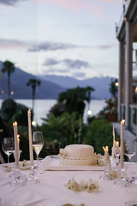 Wedding cake table with a simple white wedding cake, taper candles and crystal candlesticks on a balcony overlooking lake and mountains at dusk