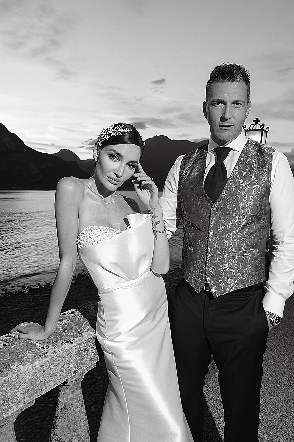 Couple portrait of bride in strapless gown with crystal hairpiece and groom in waistcoat by stone railing at a sunset lake promenade with mountains