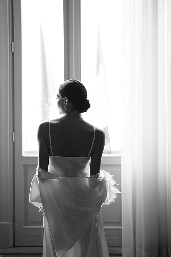 Bridal portrait of a bride in a spaghetti strap wedding dress with earrings and a low bun, standing by sheer curtains in window light