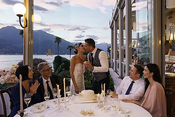 Wedding cake cutting as bride in strapless gown and groom in vest slice a two-tier cake on a terrace with lake and mountains at sunset