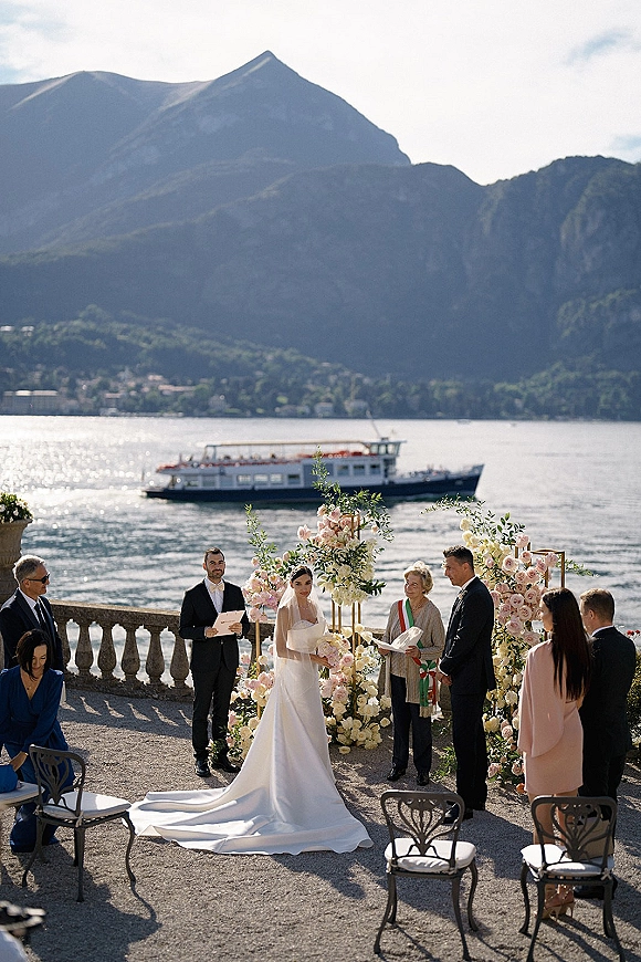 Ceremony moment at an outdoor lakeside wedding ceremony with bride and groom under a pastel rose arch on a stone terrace by mountains