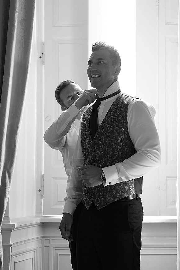 Groom getting ready as groomsmen help adjust his tie, showing patterned waistcoat, cufflinks and wristwatch in bright window light indoors