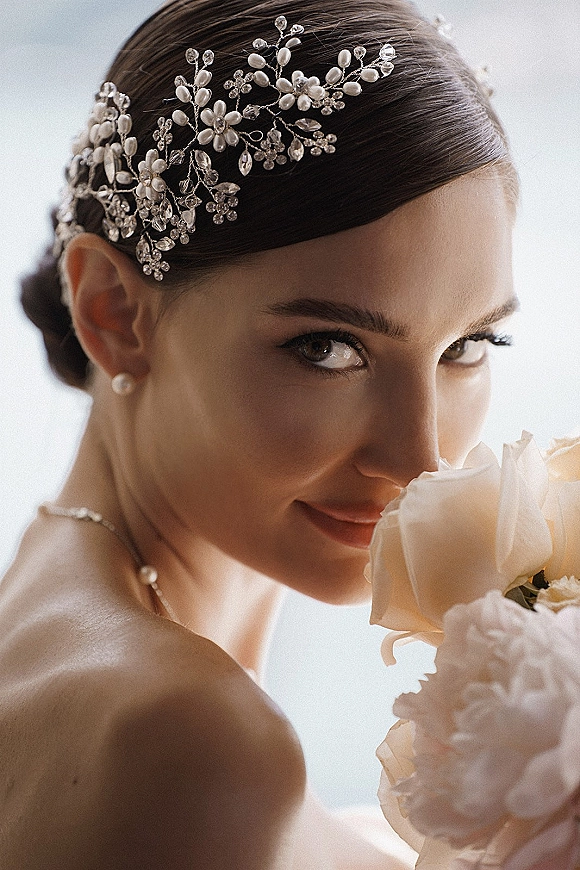 Bridal portrait of a bride with a bridal hair vine in a sleek low bun, holding an all-white rose and peony bouquet in soft sky light