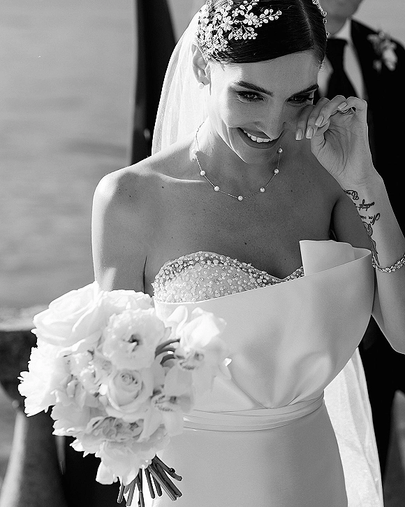 Bridal portrait in black and white of a bride wiping a tear, holding a white rose bouquet in a beaded strapless dress, groom blurred behind