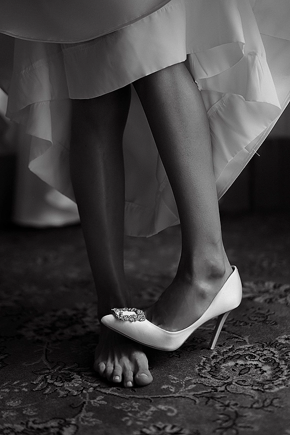 Bridal shoes with jeweled buckle as a bride in a bridal robe slips into white stiletto heels on a patterned rug indoors