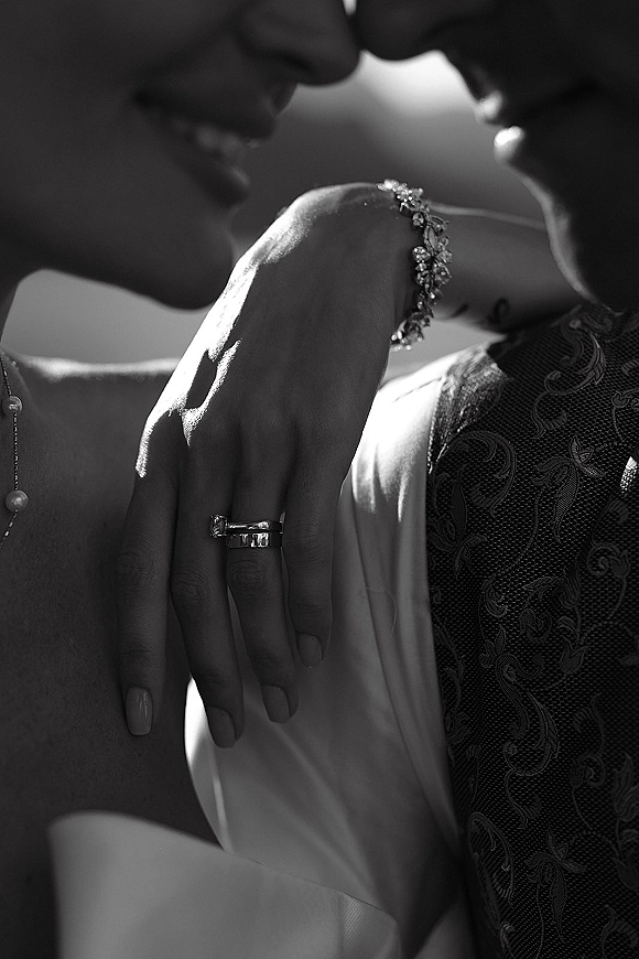 Wedding couple portrait in black and white, close up embrace with bride’s hand on groom’s chest showing stacked rings against a dark backdrop