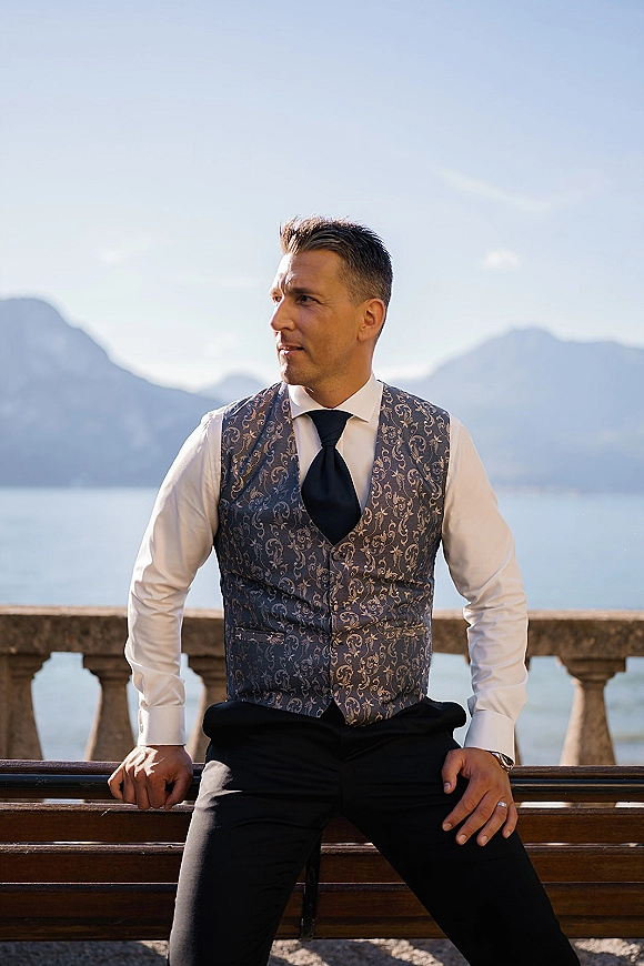 Groom portrait in a blue patterned waistcoat, leaning on a stone balustrade with a lake and mountains under a clear blue sky behind