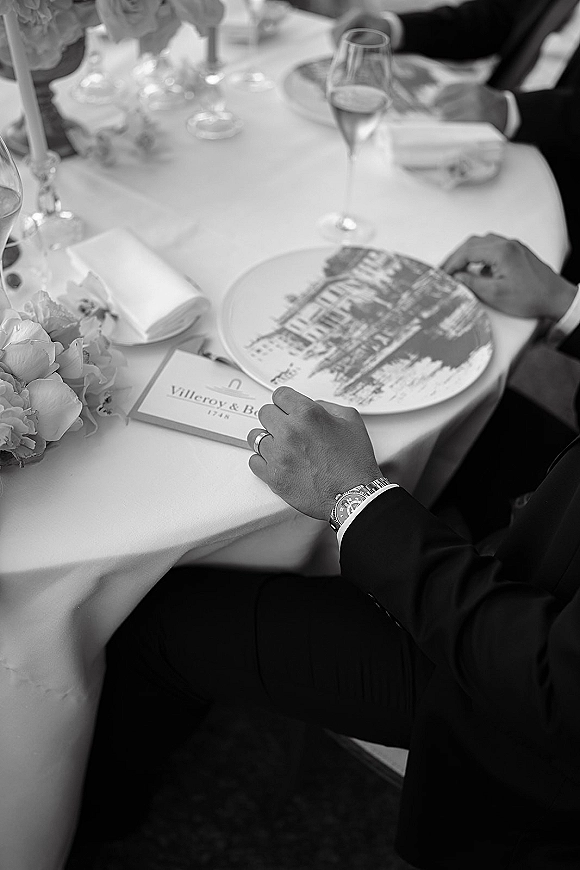 Wedding reception place setting with a custom printed charger plate, folded napkin and calligraphy place card beside glasses and taper candle at a guest table
