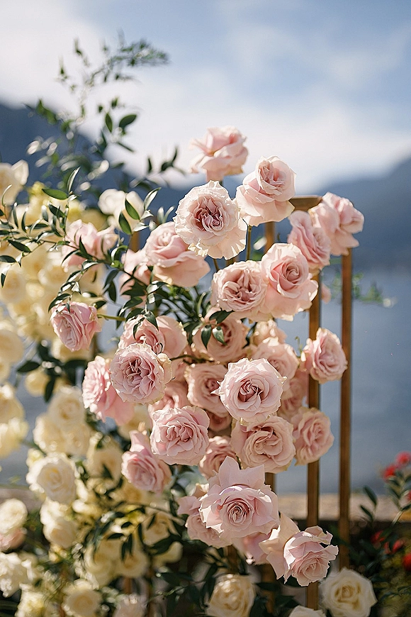 Wedding floral arch with blush roses and greenery on a gold metal frame, set against mountains, blue sky, and distant water