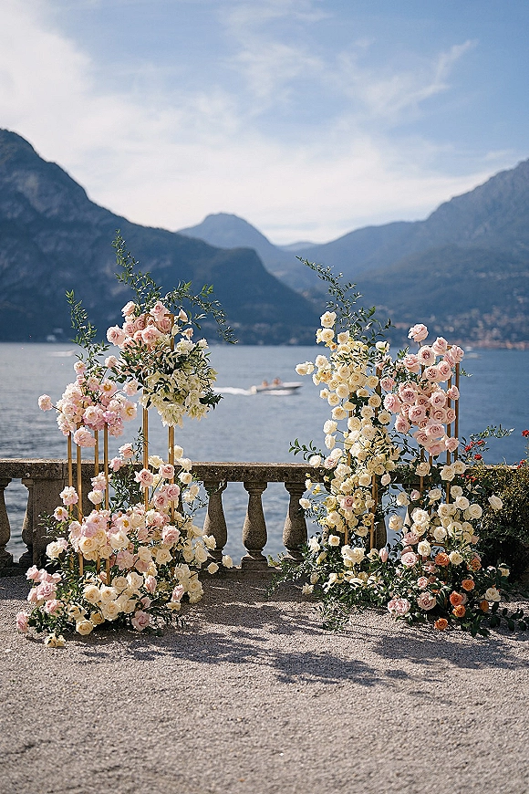 Wedding ceremony backdrop with rose florals on gold stands and greenery, set on a stone terrace overlooking a lake and mountains