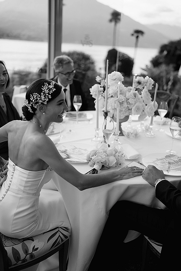 Wedding reception moment of bride at reception table holding groom’s hand, showing rings beside taper candles and florals with lake and mountains beyond windows
