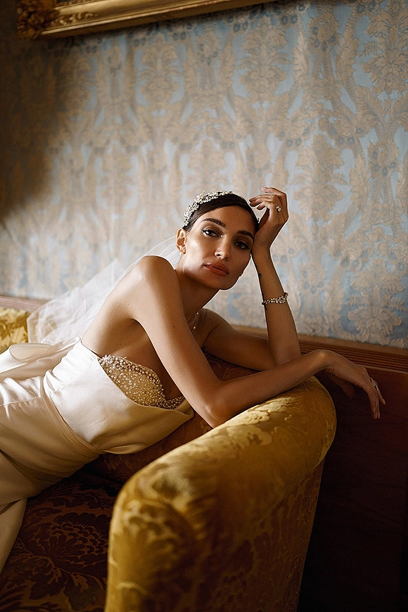 Bridal portrait of a bride reclining on a vintage sofa in a strapless beaded gown with veil and tiara against patterned wallpaper