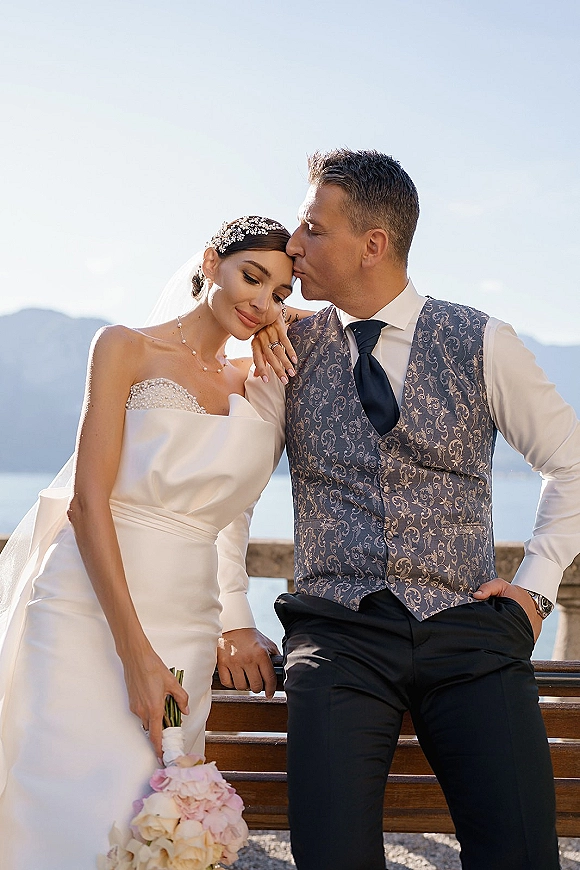 Couple portrait of bride and groom kiss, his lips on her forehead, by a lake with mountains behind, her veil flowing over a stone railing