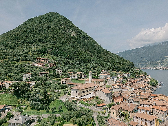 Lakeside village aerial view of terracotta roof tiles and a church bell tower above a marina, with mountains and lake below